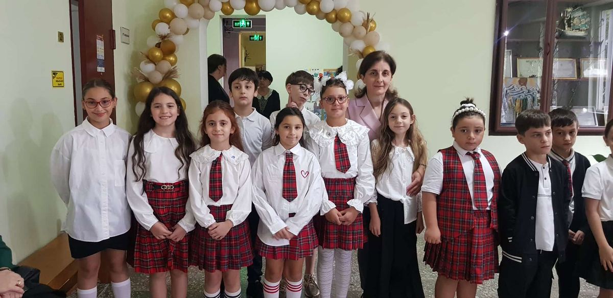 დაწყებითი კლასის მოსწავლეები სასკოლო ფორმით, მასწავლებელთან ერთად ბუშტების თაღთან